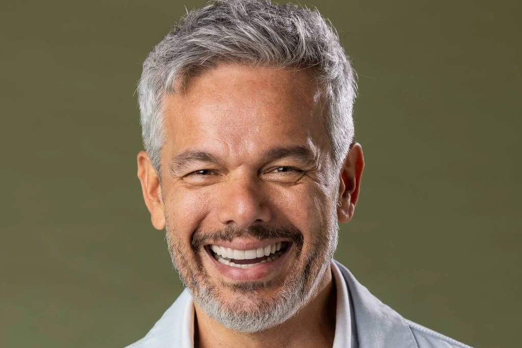 Homem de meia-idade com cabelo grisalho curto e barba aparada sorri diretamente para a câmera. Ele veste camisa branca aberta sob jaqueta clara, com fundo verde uniforme.Otaviano Costa, participante do Dança dos Famosos 2026 - Beatriz Damy/Globo